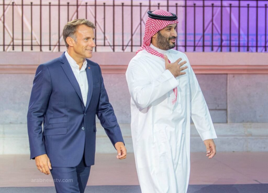 Two men walk side by side; one in a navy suit, the other in a white kandura and red‑and‑white ghutra, smiling outdoors.