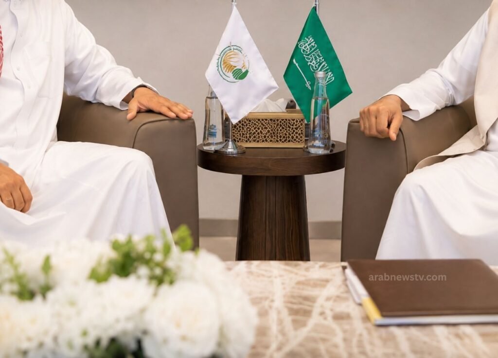 - Arab News TV Two men in white thobes sit on armchairs facing each other, with a round table, two flags, water bottles, and a tissue box between them.